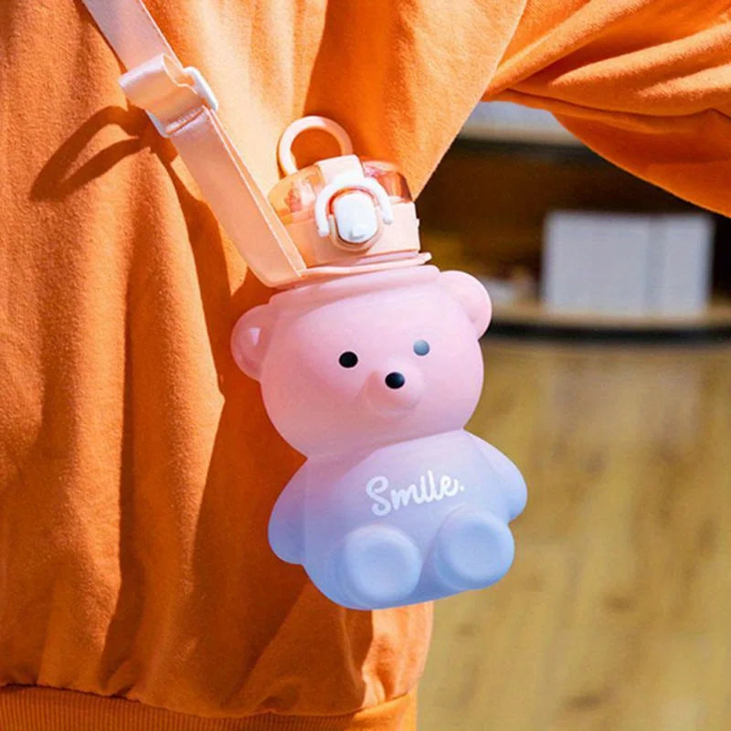 A pink bear-shaped water bottle with a straw and strap is being held by an orange-colored sleeve.