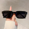 A hand holds a pair of black rectangular sunglasses against a light background.