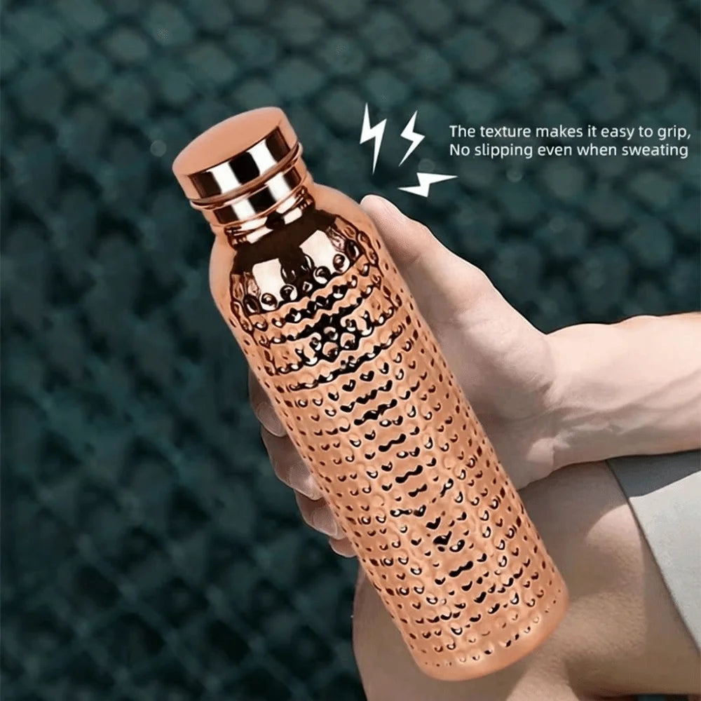 A hand holds an insulated copper water bottle with a textured anti-slip grip against a dark background.