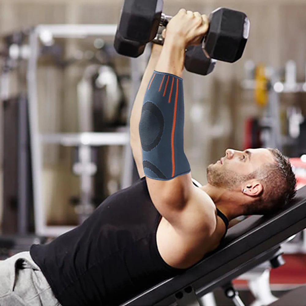 A man wearing a black shirt and an elbow support compression sleeve lifts dumbbells in a gym setting.