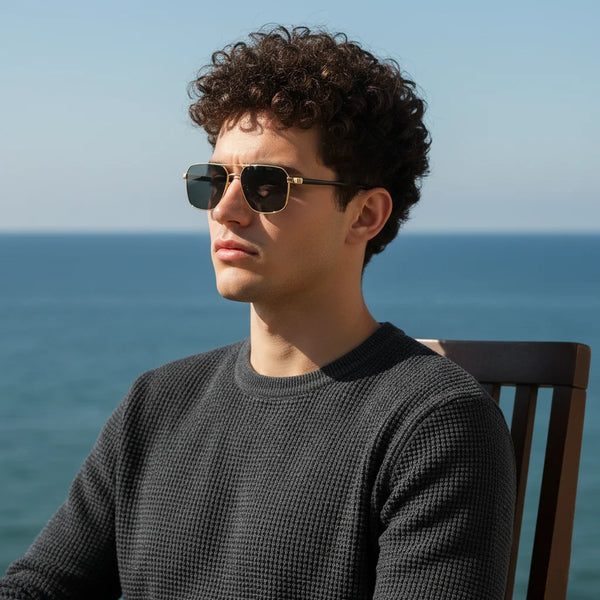 A man wearing classic square aviator sunglasses outdoors looking towards the sea.