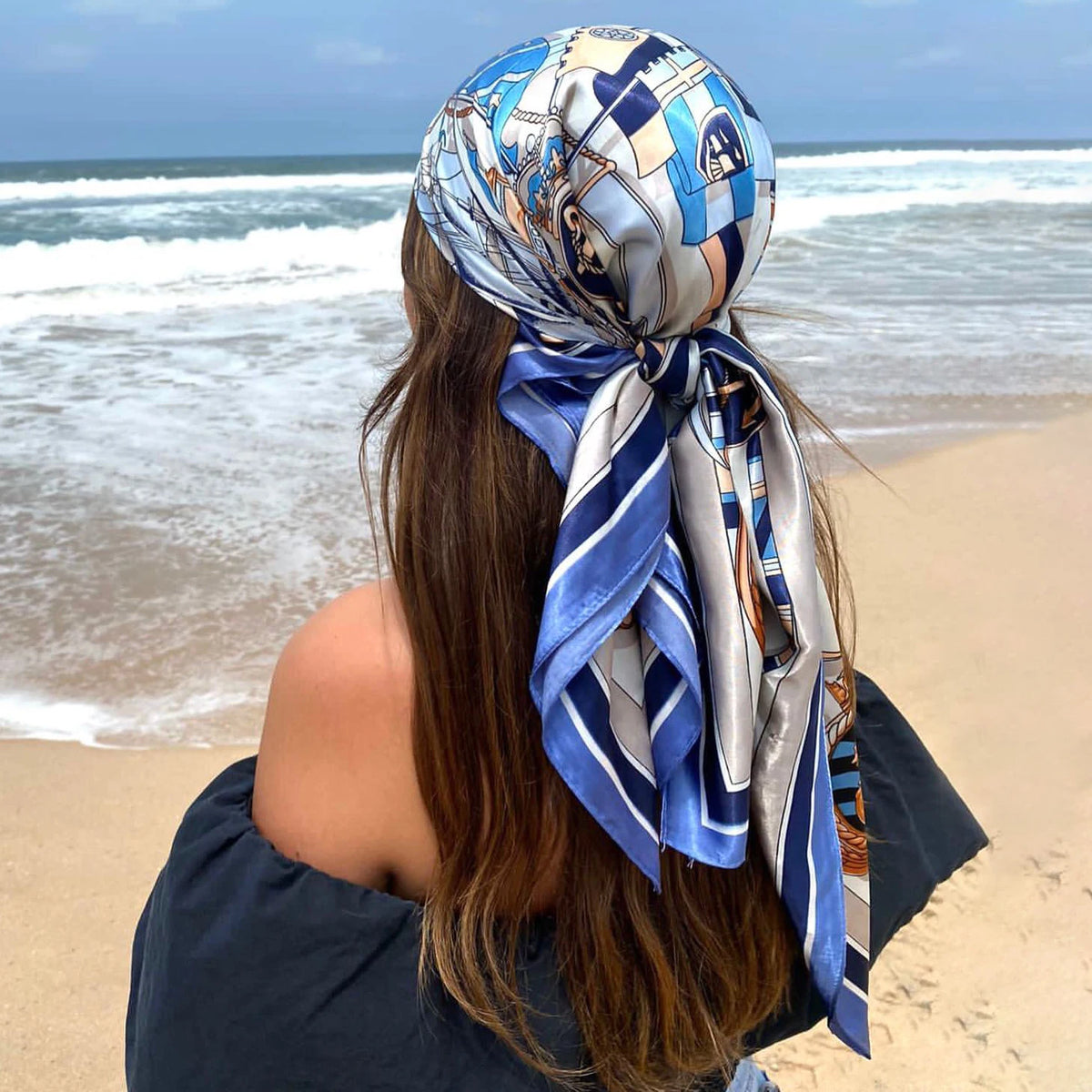 A woman with long brown hair wearing an elegant blue and silver satin square scarf tied around her hair by the ocean.