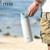 Une main tient une tasse thermos en acier inoxydable isolé blanc et bleu clair, à l'extérieur sur une plage de sable.