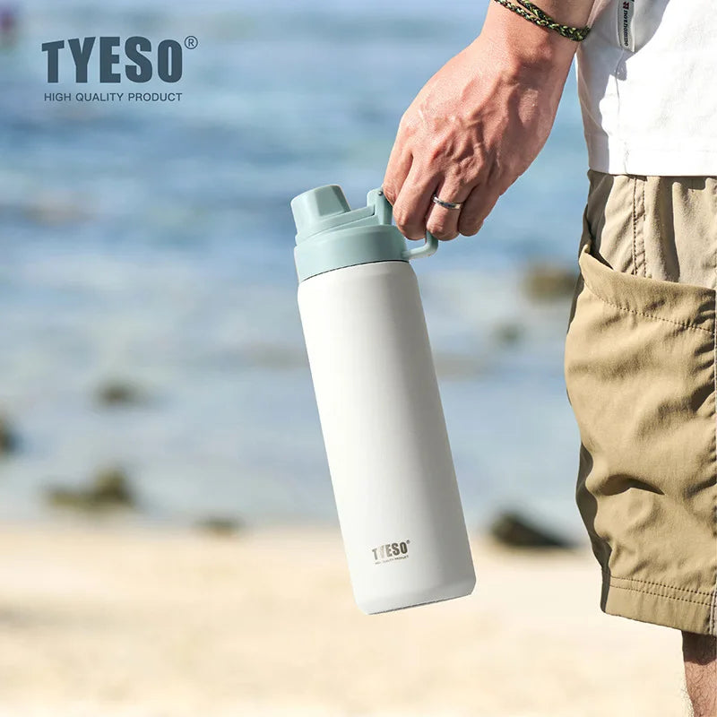 Une main tient une tasse thermos en acier inoxydable isolé blanc et bleu clair, à l'extérieur sur une plage de sable.