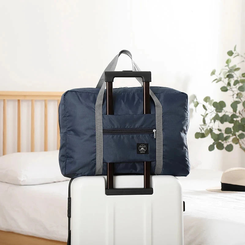 A navy blue foldable travel duffel bag with gray straps is placed on a white suitcase next to a bed.