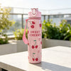 A pink reusable water bottle with a strawberry design and a pink strap, sitting on a white table outdoors.
