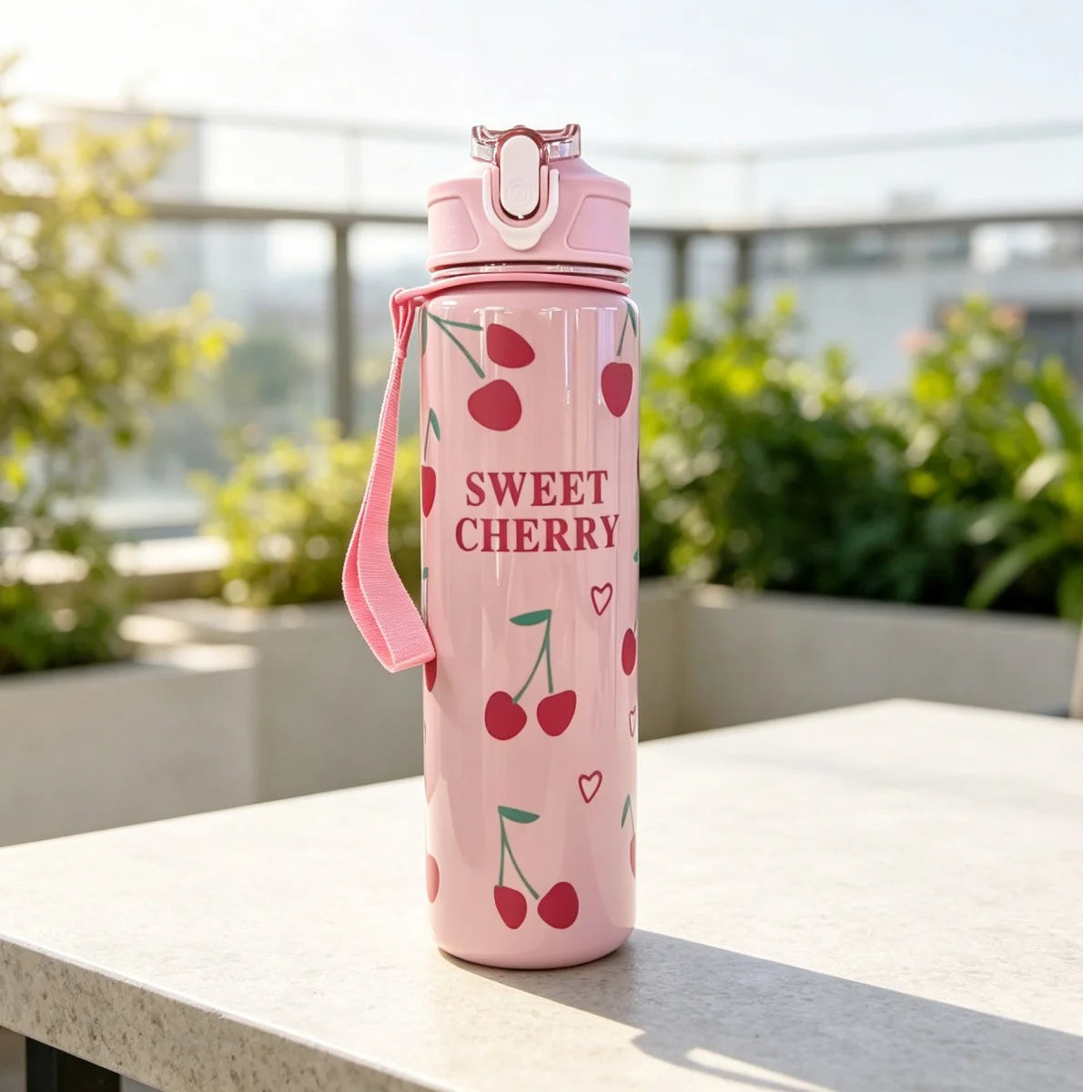 A pink reusable water bottle with a strawberry design and a pink strap, sitting on a white table outdoors.