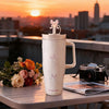A white insulated thermos mug with a pink bow on top sits on a table against a sunset cityscape.