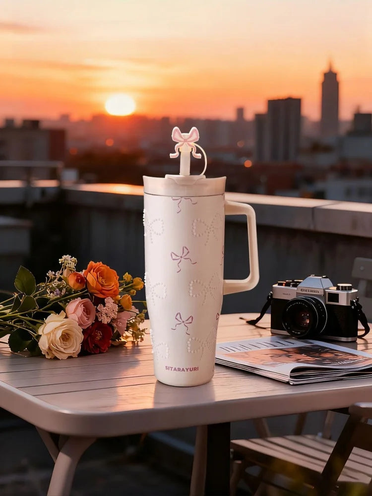A white insulated thermos mug with a pink bow on top sits on a table against a sunset cityscape.