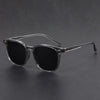 A pair of clear-framed, black sunglasses with a thin metal arm resting on a dark surface.
