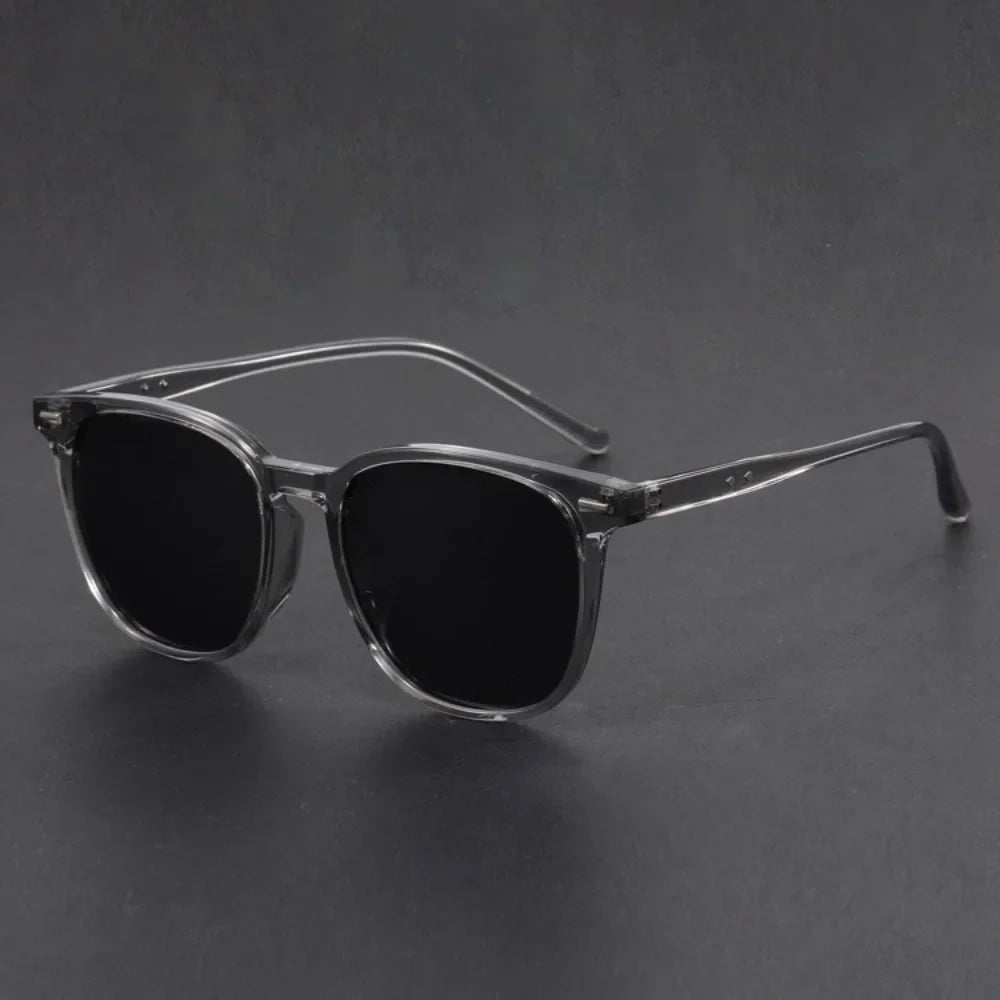 A pair of clear-framed, black sunglasses with a thin metal arm resting on a dark surface.