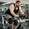 Muskulöser Mann in schwarzem Tanktop und Kniekompressionsärmeln fährt ein stationäres Fahrrad im Fitnessstudio.