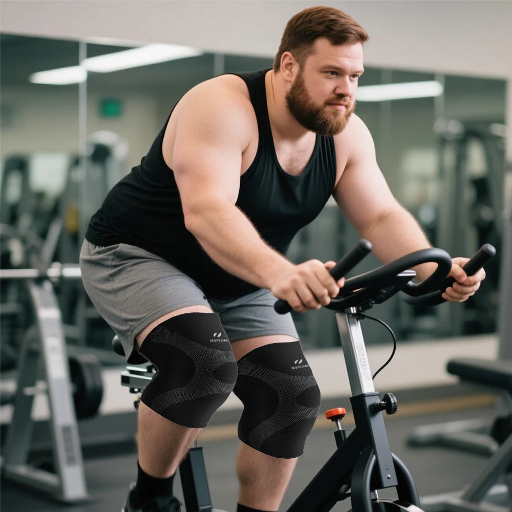 Muskulöser Mann in schwarzem Tanktop und Kniekompressionsärmeln fährt ein stationäres Fahrrad im Fitnessstudio.