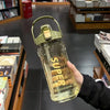 A hand holds a clear glass insulated sports water bottle with a gold-colored cap and straw against a dark background.