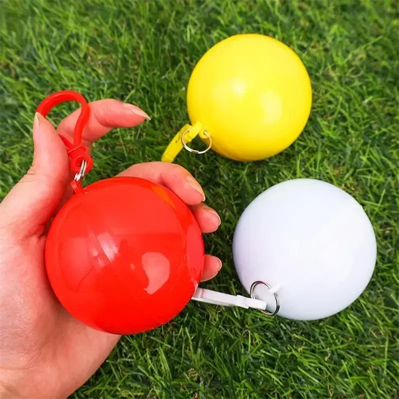 A hand holds a red portable disposable rain poncho and a yellow inflatable ball on green grass.
