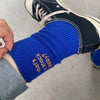 A close-up of a woman's ankle wearing bright blue knitted socks with gold embroidery, paired with black and white sneakers.