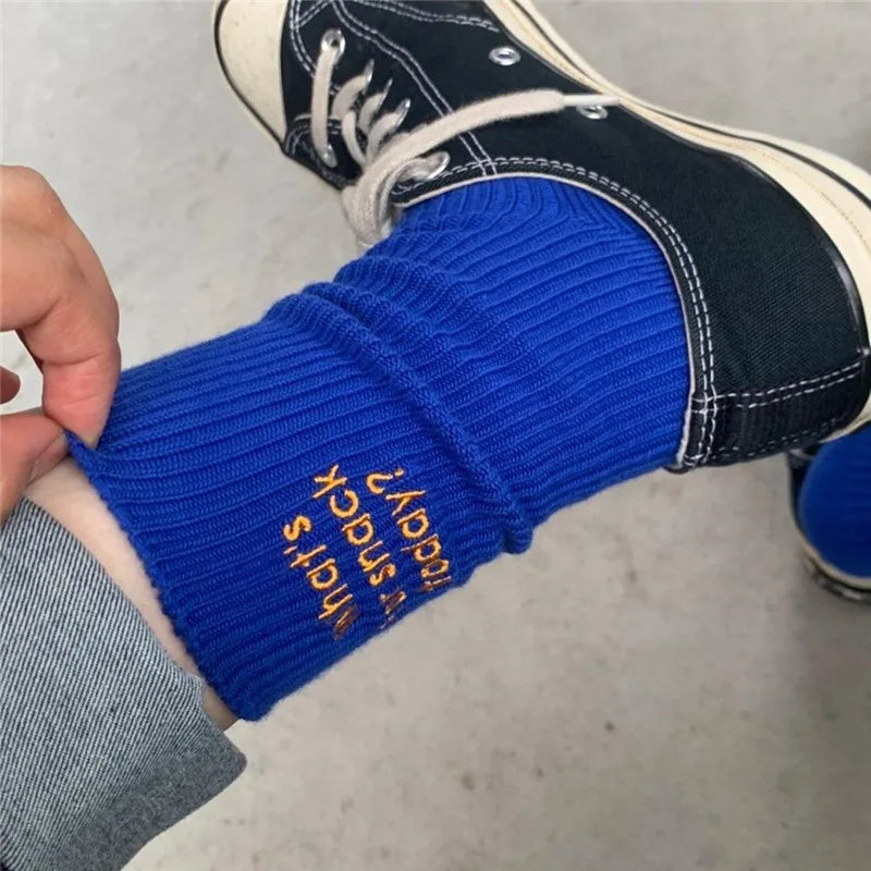 A close-up of a woman's ankle wearing bright blue knitted socks with gold embroidery, paired with black and white sneakers.