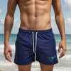 A muscular man wearing navy blue quick-drying swim shorts standing near the ocean.