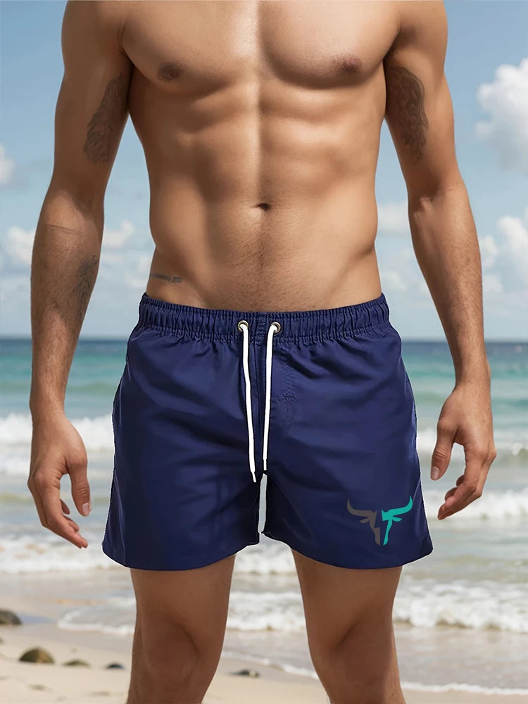 A muscular man wearing navy blue quick-drying swim shorts standing near the ocean.