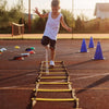 A young girl runs across a yellow agility ladder on an orange sports field at sunset.