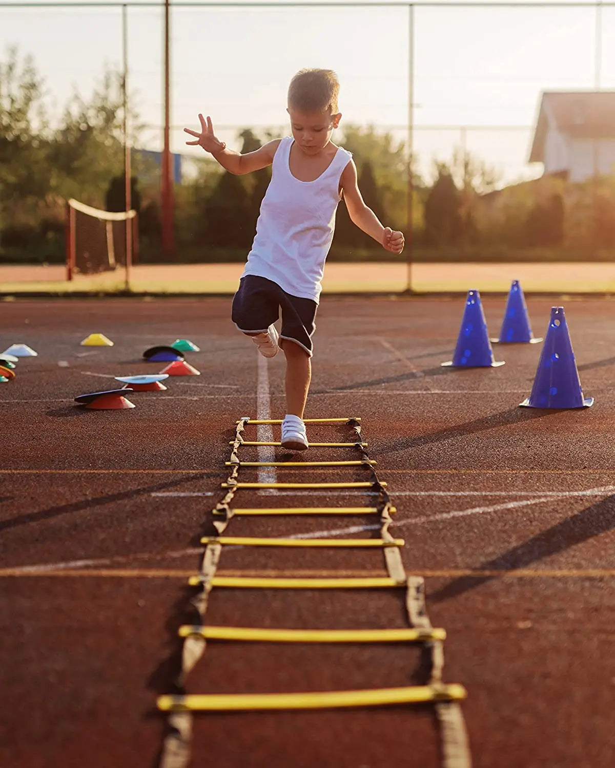 A young girl runs across a yellow agility ladder on an orange sports field at sunset.