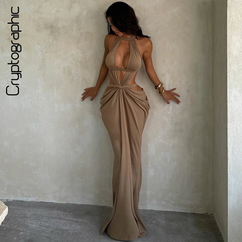 A woman wearing a floor-length, draped, beige maxi dress with a cut-out back stands against a light wall.