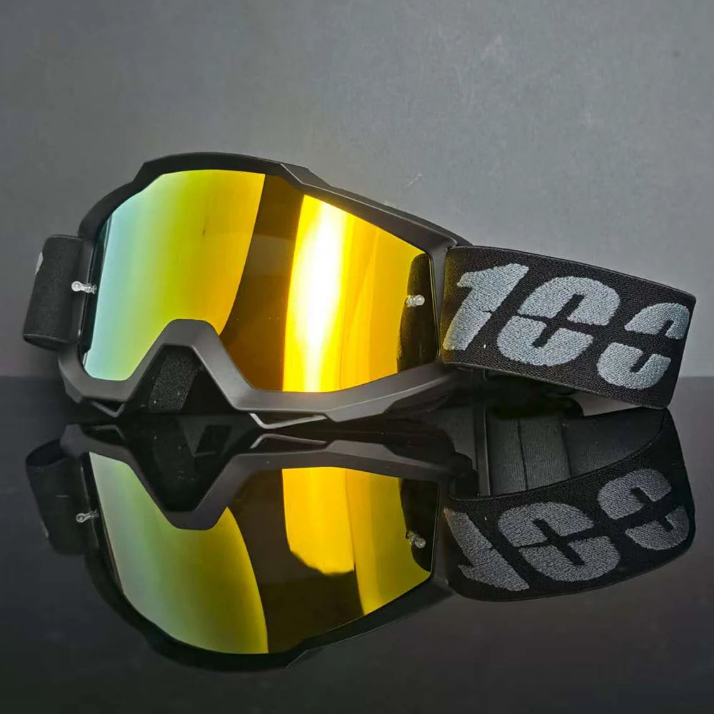 Un par de gafas de motocross negras con lentes tintados en amarillo y una correa negra, reflejadas en una superficie oscura.