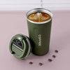 A dark green insulated thermos bottle with a lid is shown next to a cup of latte art on a pink surface.