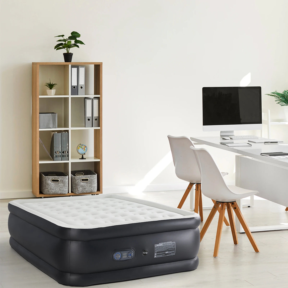 A black air mattress is placed on the floor next to a wooden shelf and a white desk.