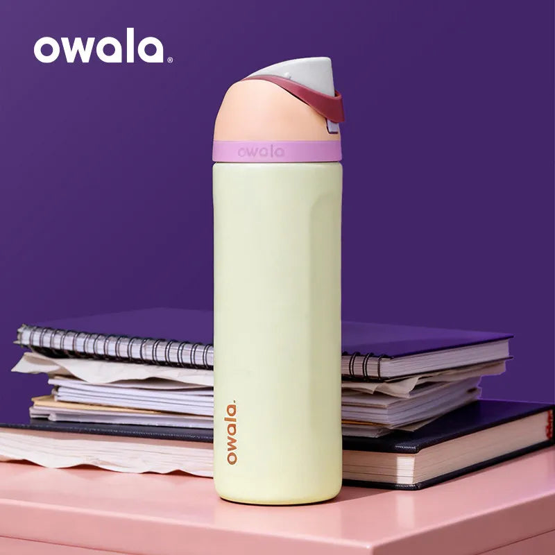 A light yellow Owala insulated tumbler with a pink lid is shown standing on a pink surface next to stacked notebooks.