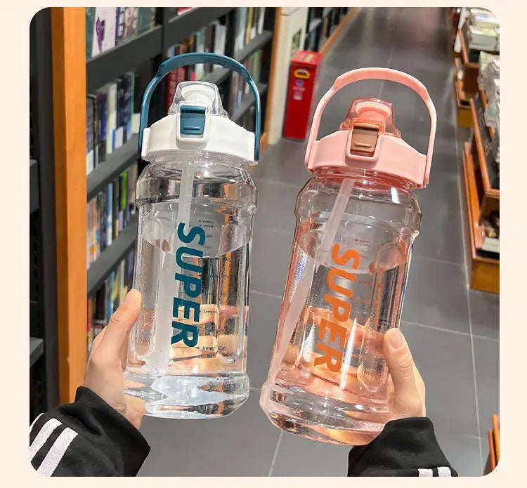 A hand holds a clear insulated sports water bottle with a pink lid and straw against a blurred background of bookshelves.