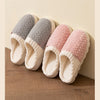 A set of four plush fleece slippers in gray, pink, and cream colors resting on a wooden floor.