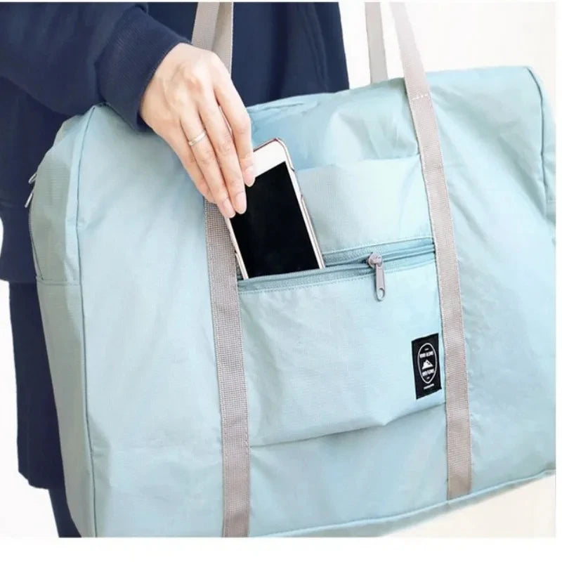 A hand is placing a smartphone into the pocket of a light blue foldable travel duffel bag.