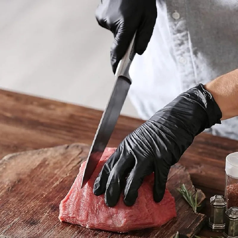 Une main portant un gant en nitrile noir utilise un couteau pour couper de la viande rouge crue sur une planche à découper en