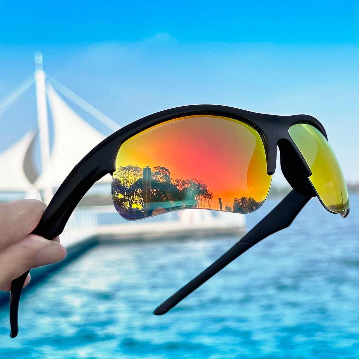 A hand holds a pair of black sports sunglasses with yellow-tinted lenses against a bright blue sky and water.