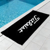 A black microfiber beach towel is laid on a tiled patio next to a swimming pool.
