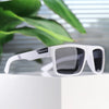 White polarized square sunglasses resting on a white surface with green foliage in the background