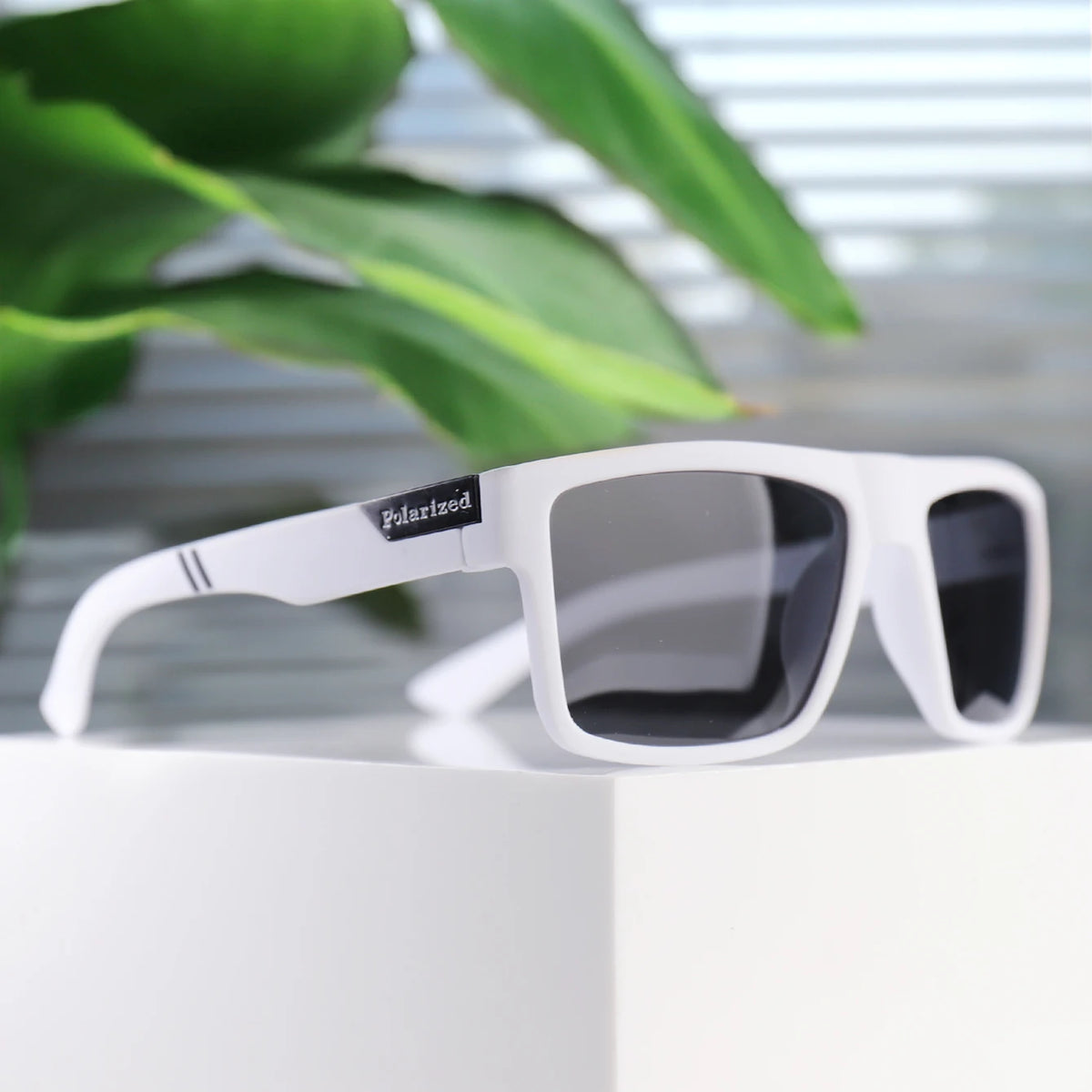 White polarized square sunglasses resting on a white surface with green foliage in the background