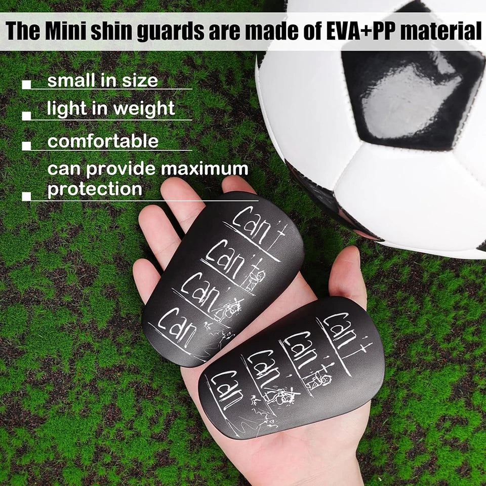 A hand holds black mini shin guards made of EVA+PP material next to a soccer ball.