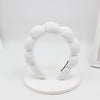 A white, fluffy sponge terry cloth headband for hair styling displayed on a white circular stand.