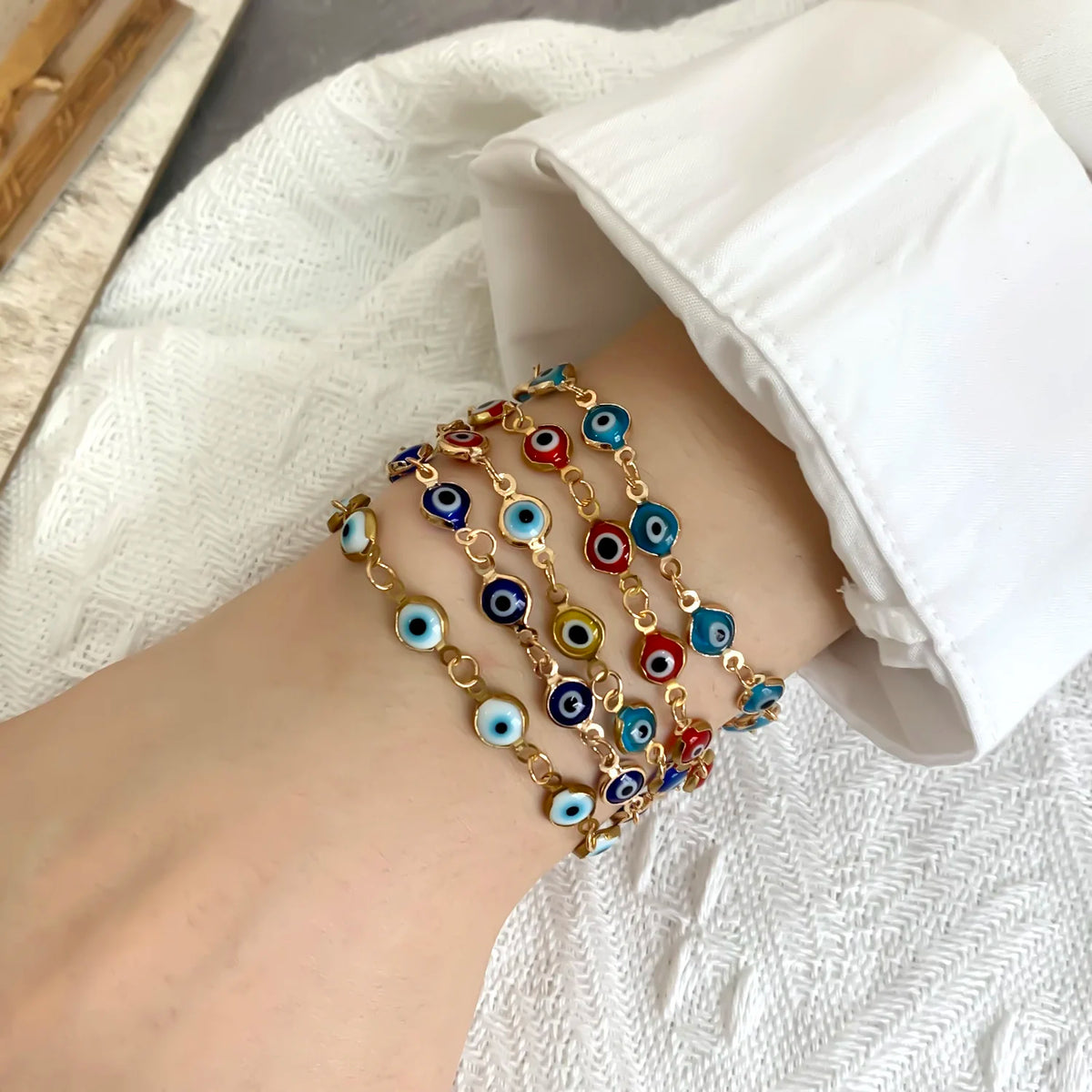 A close-up of a wrist wearing multiple gold charm bracelets featuring blue and multicolored evil eye beads.