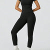 Eine Frau trägt ein schwarzes zwei-teiliges Yoga-Set mit hohen Leggings und einem kurzärmeligen Oberteil.