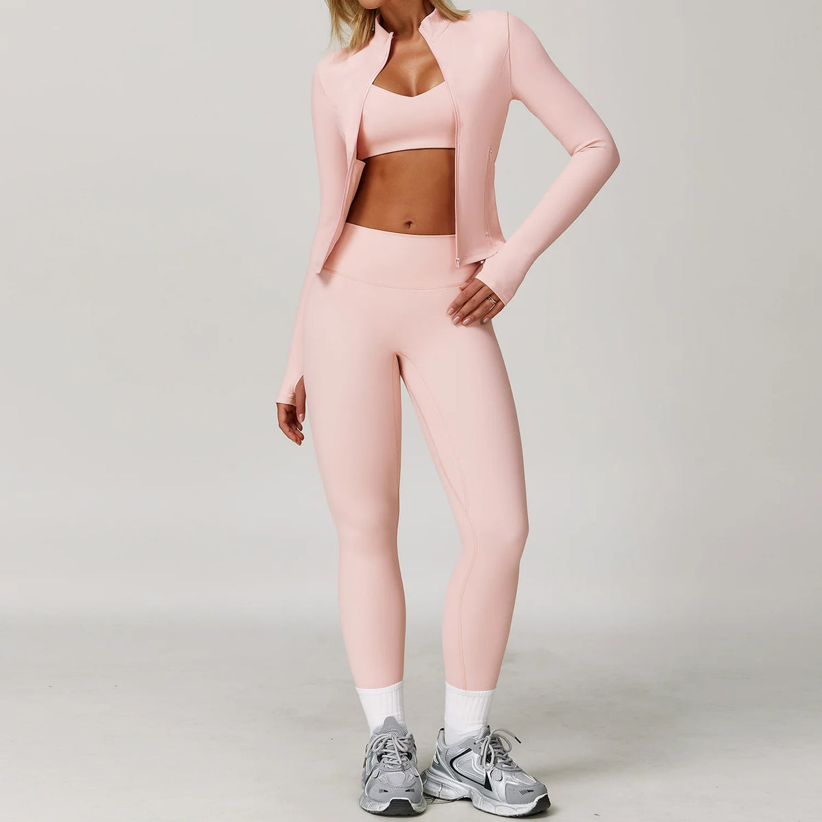A woman models a pink 3-piece yoga sports set consisting of a long-sleeved cropped jacket, sports bra, and leggings.