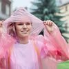 A young woman smiling while holding a sheer pink disposable rain poncho over her head outdoors.