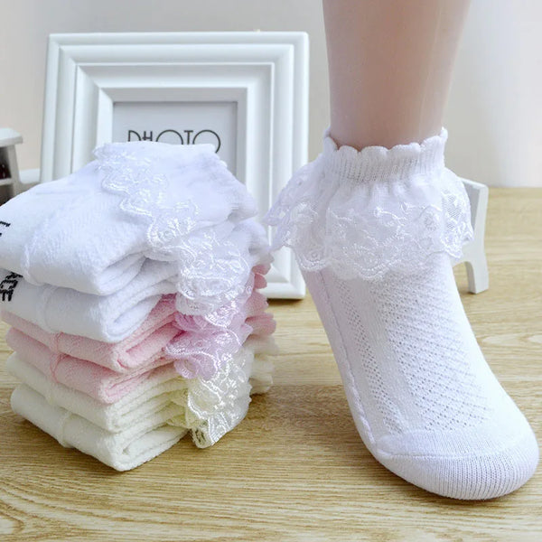 Un par de calcetines de encaje con volantes blancos para niñas, junto a una pila de calcetines rosa y blanco.