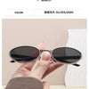 A hand holds a pair of silver/gray retro oval sunglasses against a white background.