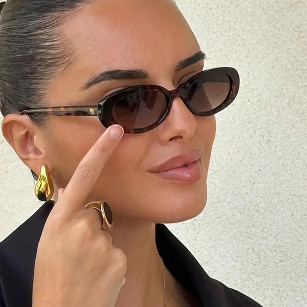 A woman wearing dark brown retro oval sunglasses with gold earrings and a black blazer.