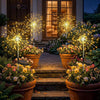 Luces de hadas solares iluminando un camino de piedra de jardín con flores en macetas y fuegos artificiales.