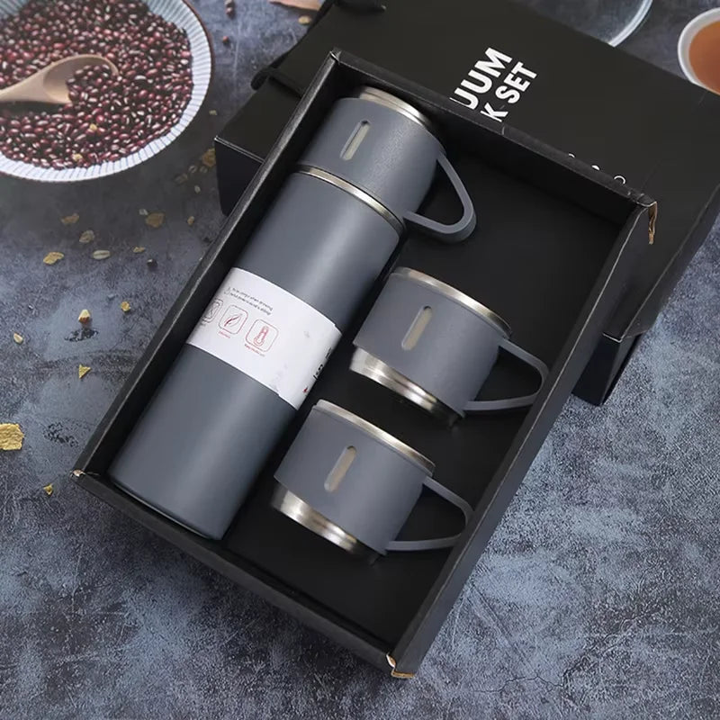 A black box contains a set of four grey stainless steel vacuum flasks and a measuring spoon on a dark surface.