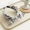 A black and white paisley bandana headband tied in a bow, displayed on a light tray next to a white cup.